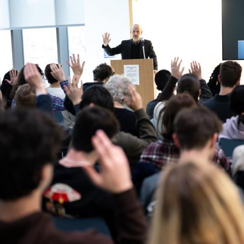Peter Adamson speaks at a podium while many in the audience raise their hands.